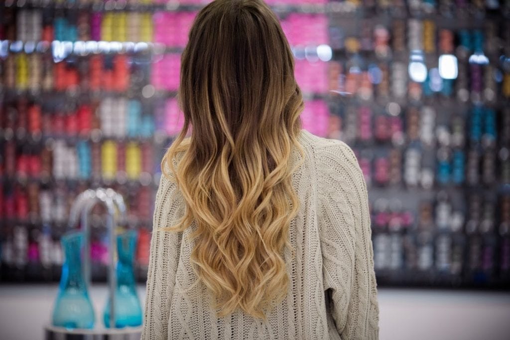 Человек с длинными волнистыми светлыми волосами в омбре, одетый в свитер кремового цвета, стоит перед красочной, размытой витриной в салоне или магазине в Ногинске.